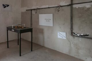 Ein Industrieraum mit freiliegenden Rohren, zwei weißen Wandtafeln, einem schwarzen Ausstellungstisch mit Gegenständen unter Glas und einem weißen Objekt auf einem Rohrregal.