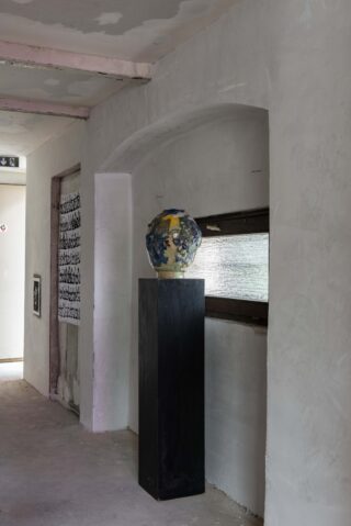 Eine Keramikkopfskulptur steht auf einem hohen schwarzen Sockel in einem minimalistischen Flur mit strukturierten weißen Wänden und einem schmalen Fenster.