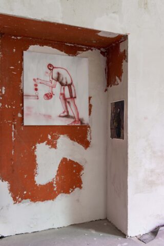 An einer Wand mit abblätternder Farbe hängt das Gemälde einer Person, die Tischtennis spielt. Rechts daneben ist ein kleineres Foto oder ein Druck zu sehen.