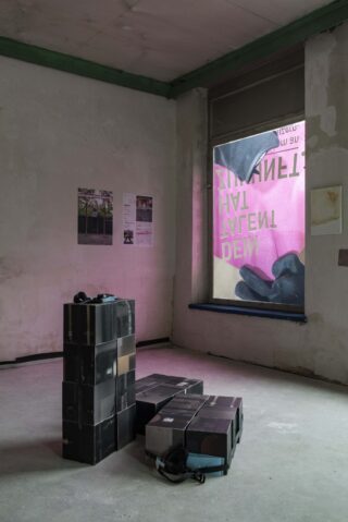 Ein minimalistischer Raum mit einem Stapel schwarzer Kästen, einem Headset, verblassten Postern an der Wand und einem großen Fenster, vor dem ein umgekehrtes rosa Schild zu sehen ist.