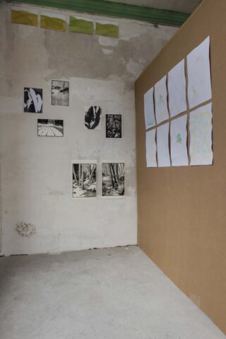 In einer kargen Galerieecke hängen Schwarzweißfotos an einer grauen Wand und mehrere abstrakte Zeichnungen an einer braunen Wand. Der Boden besteht aus nacktem Beton.