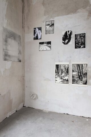 In einer Ecke eines Raumes mit abblätternder Farbe sind acht abstrakte und landschaftliche Kunstwerke in Schwarzweiß an der Wand über einem Betonboden angebracht.