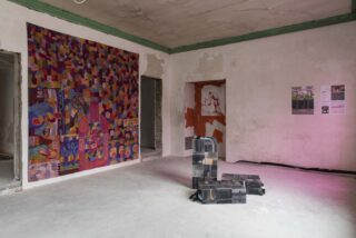 Eine Kunstinstallation in einem kahlen Raum besteht aus einem farbenfrohen abstrakten Wandteppich, einer kastenförmigen Skulptur, Graffiti und zwei Postern auf einer rauen, unbearbeiteten Wand.