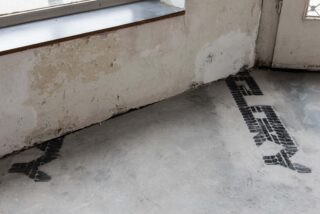 Auf einem Betonboden in der Nähe einer Wand und einer Tür sind schwarze Reifenspuren zu sehen. Darüber blättert die Wandfarbe ab und es gibt ein Fenster.