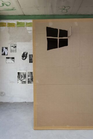 In einem Raum steht eine große Pappwand mit Fensterausschnitt, im Hintergrund sind auf einer weißen Wand Schwarz-Weiß-Fotos zu sehen.