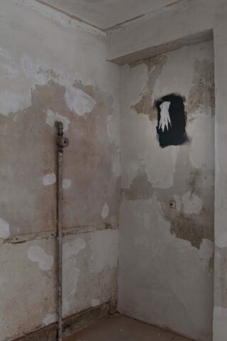 Eine abgenutzte, abblätternde Wand mit freiliegenden Rohrleitungen und einer Schablone aus weißen Handschuhen, die auf ein schwarzes Rechteck gemalt ist.