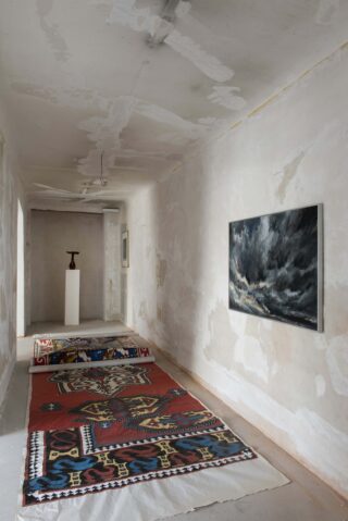 Ein schmaler Raum mit unfertigen Wänden, an denen ein großer Teppich, ein Sockel mit einer kleinen Skulptur und zwei gerahmte Kunstwerke hängen, darunter ein Gemälde eines Gewitterhimmels.