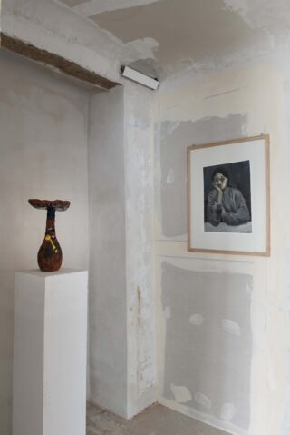 Eine Keramikvase auf einem weißen Sockel steht neben einem gerahmten Schwarzweißporträt an einer Wand mit unbehandeltem Putz und sichtbaren Flecken.