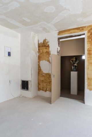 Ein teilweise renovierter Raum mit freiliegendem Putz, einer offenen Tür, die in einen dunklen Raum führt, und einer Skulptur auf einem Sockel, die im nächsten Raum sichtbar ist.