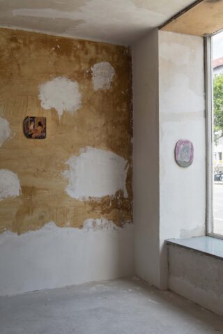 Ein teilweise renovierter Raum mit geflickten, unfertigen Wänden, zwei kleinen hängenden Gemälden und einem großen Fenster mit Blick auf eine Straße.