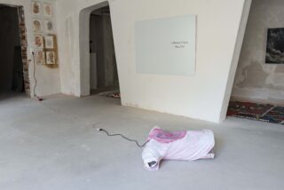 Auf dem nackten Betonboden liegt ein rosa gepolstertes Objekt, das mit einem schwarzen Kabel angeschlossen ist; an den minimalistisch weißen Wänden sind verschiedene gerahmte Kunstwerke zu sehen.
