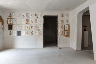 Ein kahler Raum mit Wänden und Boden aus unbearbeitetem Gips. An einer Wand sind mehrere Kunstdrucke in einem Raster angeordnet, im angrenzenden Raum steht eine Skulptur auf einem Sockel.