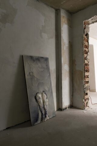 Ein strukturiertes Gemälde lehnt an einer kahlen, unbehandelten Wand in einem Raum mit freiliegenden Ziegel- und Betonoberflächen.