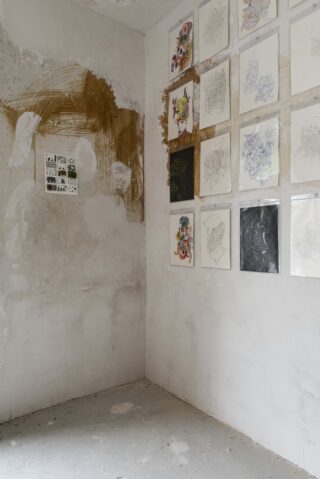 In einer Ecke eines kahlen, unfertigen Raums sind an einer Wand mehrere Zeichnungen und Skizzen angebracht, der umgebende Putz weist Schmierer und braune Flecken auf.