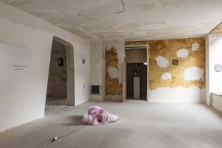 Ein spärlich möblierter, unfertiger Raum mit fleckigen Wänden, einem kleinen Kunstwerk an einer Wand und einem rosa-weißen Deckenbündel auf dem Boden neben einem Stromkabel.