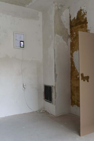 Eine kleine elektronische Anzeige und Papier sind an einer rauen, teilweise gestrichenen Wand in einem unfertigen Raum mit freiliegenden Kabeln und einem schräg stehenden Stück Pappe angebracht.
