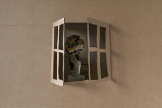 Eine Pappwand mit einem ausgeschnittenen Fenster öffnet sich und gibt den Blick auf eine darin ausgestellte Keramikskulptur frei.