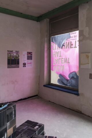 Ein minimalistisch eingerichteter Raum mit kahlen Wänden, zwei Postern, gestapelten schwarzen Kisten und einem Fenster, das mit einem rosa getönten, durchscheinenden Banner mit umgekehrtem Text bedeckt ist.