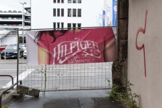 Auf einem städtischen Parkplatz ist an einem Zaun neben einer Mauer mit rotem Graffiti ein Banner angebracht, das eine Person in einem rosa „Hilfiger Denim“-Hemd zeigt.