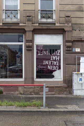 Eine Gebäudefassade mit zwei großen Fenstern; in einem Fenster hängt ein Plakat mit umgekehrtem deutschen Text. Autos und Gebäude spiegeln sich im Glas.