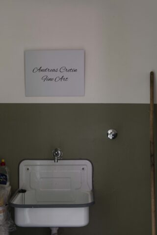 Ein weißes Waschbecken an einer zweifarbigen Wand, darüber ein Schild mit der Aufschrift „Andreas Cretin Fine Art“ in Schreibschrift und ein Moppgriff, der an der Wand lehnt.