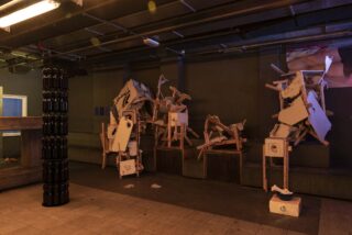 Ein schwach beleuchteter Raum mit abstrakten Skulpturen aus zerbrochenem Holz und weißen Tafeln auf Bänken, neben einem vertikalen Stapel schwarzer zylindrischer Objekte.