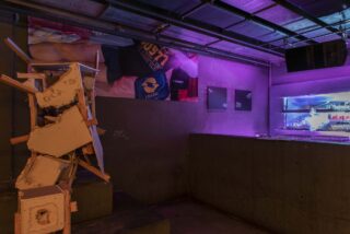 Schwach beleuchteter Raum mit violetter Beleuchtung, einer zerbrochenen Skulptur aus Schutt, Postern an einer grauen Wand und einer neonbeleuchteten Vitrine auf der rechten Seite.