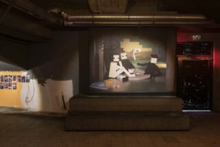 Ein schwach beleuchteter Raum mit Industrierohren, einem digital anmutenden Gemälde abstrakter Figuren auf einer Bühne, einer Fotocollage an einer Wand und einem dunklen Türrahmen auf der rechten Seite.