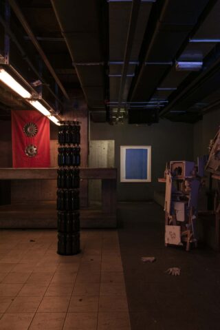 Schwach beleuchteter Industrieraum mit freiliegenden Rohren, einer roten Flagge mit Zahnrädern an der Wand, einer Säule aus schwarzen Gefäßen und einer unordentlichen Struktur aus gestapelten Objekten auf der rechten Seite.