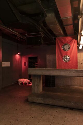 Ein Betonraum mit freiliegenden Rohren, einem roten Licht, einem roten Tuch mit zwei Radkappen an der Wand und einem Bündel weißen Stoffs auf dem gefliesten Boden.