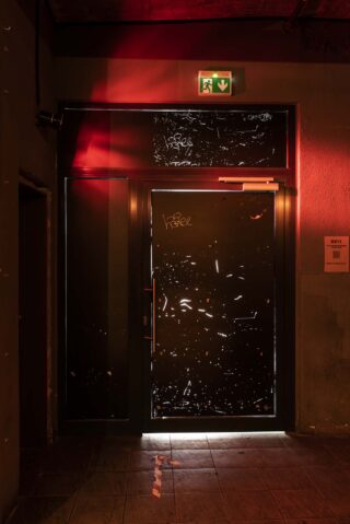 Eine schwach beleuchtete Ausgangstür mit Graffiti wird durch rotes Licht beleuchtet, darüber befindet sich ein grünes Notausgangsschild und unter der Tür ist ein Lichtstrahl sichtbar.