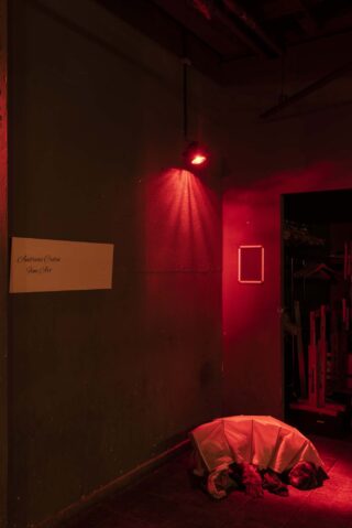 Eine schwach beleuchtete Ecke mit einem roten Licht darüber, ein weißes Laken bedeckt einen Gegenstand auf dem Boden und ein Schild an der Wand mit der Aufschrift „Montana Cueto Fine Art“.