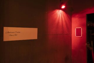 Ein schwach beleuchteter Raum mit rotem Licht, einem leeren Bilderrahmen an einer Wand und einem Schild mit der Aufschrift „Andreas Creten Fine Art“ an einer anderen Wand.