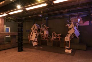 Ein schwach beleuchteter Raum mit abstrakten Skulpturen aus gestapelten Möbeln und Pappe, schwarzen zylindrischen Objekten, gefliestem Boden und freiliegenden Deckenrohren.