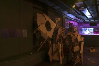 Schwach beleuchteter Raum mit abstrakten Skulpturen aus Holz und Trockenbau, drei strukturierten Paneelen an der Wand und einer Bar mit Flaschen und Neonlichtern im Hintergrund.