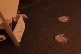 Zwei geformte Handabdrücke liegen auf einem dunklen Fliesenboden neben einer teilweise zerbrochenen weißen Struktur mit freiliegenden Holzstützen.