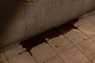 Eine dunkelrote Flüssigkeit hat sich am Fuß einer rauen Betonwand auf einem beigefarbenen Fliesenboden angesammelt und verschmiert.