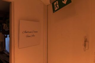 Neben einer Tür mit Klinke befindet sich ein Wandschild mit der Aufschrift „Andreas Cortin Fine Art“. Über der Tür befindet sich ein Notausgangsschild.
