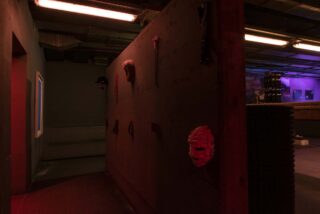 Schwach beleuchteter Raum mit dunklen Wänden, an denen verschiedene Masken und Objekte ausgestellt sind; Leuchtstoffröhren beleuchten Teile des im Industriestil gestalteten Raums.