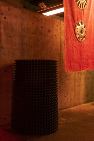 Ein zylindrisches schwarzes Objekt mit einer spitzen Oberfläche steht auf einem Betonboden neben einer Betonwand und einem hängenden roten Stoff mit kreisförmigen metallischen Verzierungen.