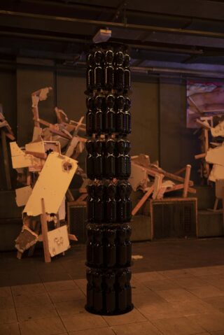 Ein hoher vertikaler Stapel identischer schwarzer Flaschen steht in einem Industrieraum, im Hintergrund sind verstreute Holztrümmer und zerbrochene Bretter zu sehen.