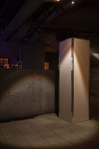 Ein hohes, rechteckiges, freistehendes Paneel mit Metallrahmen steht auf einem gefliesten Boden in einem schwach beleuchteten Industrieinterieur mit freiliegenden Rohren und Betonoberflächen.