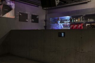 Ein Barkeeper mit Maske steht hinter einer Bartheke aus Beton in einem schwach beleuchteten Raum mit Regalen voller Flaschen und Gläser und Speisekarten an der Wand.