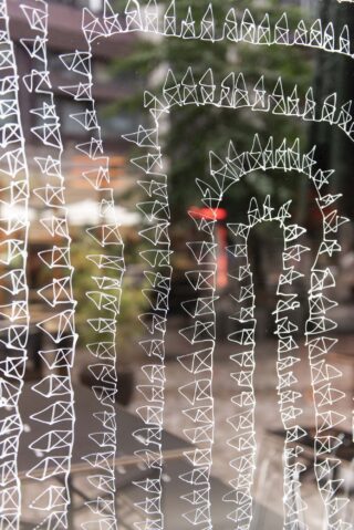 Weiße geometrische Muster, die Dreiecken und Bögen ähneln, sind auf eine transparente Oberfläche gezeichnet, im Hintergrund ist eine Außenszene unscharf.