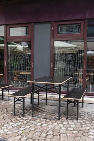 Ein leerer schwarzer Tisch mit zwei Bänken steht auf einer gepflasterten Terrasse vor einem Gebäude mit großen Fenstern und dekorativer Schrift.
