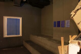 Ein schwach beleuchteter Raum mit Betonstufen, einem großen gerahmten blauen Kunstwerk an der linken Wand, drei kleineren blauen Stücken an der Rückwand und Holztrümmern im rechten Vordergrund.