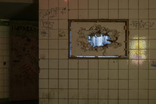 Eine geflieste Wand mit Graffiti und einem großen, unregelmäßigen Loch in der Mitte, das durch die Öffnung einen Blick auf einen anderen Raum freigibt.