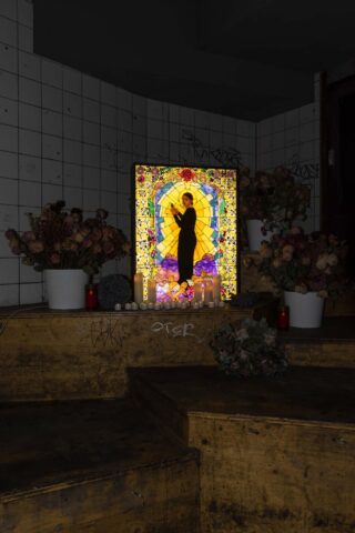 Auf abgenutzten gelben Stufen ist ein Buntglasfenster mit der Darstellung eines Heiligen beleuchtet, umgeben von getrockneten Blumen in Töpfen und brennenden Kerzen in einem gefliesten Innenraum.