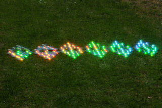 Sechs geometrische Formen aus kleinen, bunten LED-Lichtern, die in einer Reihe auf einer Rasenfläche angeordnet sind.