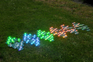 Auf dem Gras sind in Gruppen angeordnete CDs angebracht, die das Sonnenlicht reflektieren und so blaue, grüne und orange Farbtöne erzeugen.